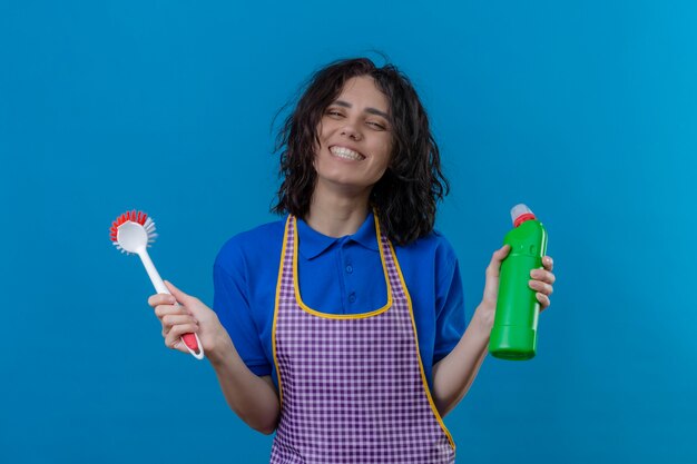 Mujer joven vistiendo delantal sosteniendo un cepillo y una botella de productos de limpieza sonriendo alegremente mirando alegre de pie sobre fondo azul.