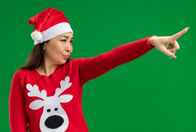 Foto gratuita mujer joven vistiendo navidad santa sombrero y suéter rojo mirando a un lado con cara seria apuntando con el dedo índice de pie sobre fondo verde