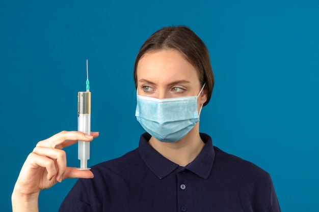 Mujer joven vistiendo polo azul en máscara médica protectora con jeringa mirándolo fijamente con cara seria de pie sobre fondo azul aislado