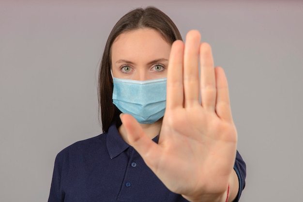 Mujer joven vistiendo polo azul en máscara médica protectora mostrando gesto de parada de mano con cara seria aislado sobre fondo gris claro
