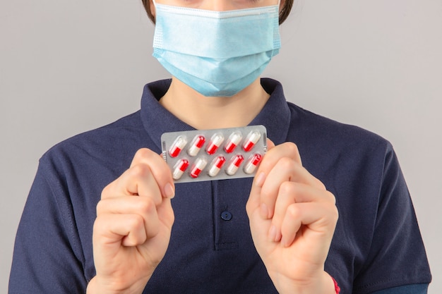 Foto gratuita mujer joven vistiendo polo azul en máscara médica protectora con pastillas de ampolla en manos sobre fondo gris claro