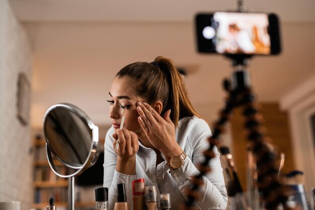 Mujer joven vlogueando sobre productos de belleza mientras aplica sombra de ojos en casa