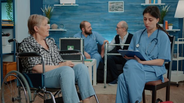 Mujer jubilada con discapacidad hablando con una enfermera para un chequeo médico en un hogar de ancianos. Asistente de atención médica que consulta a un paciente mayor sentado en silla de ruedas para obtener apoyo y asistencia.