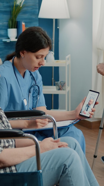 Mujer jubilada y enfermera hablando con un médico en una videollamada para telemedicina y telesalud en un hogar de ancianos. Asistente médico sosteniendo un teléfono inteligente, ayudando a un paciente discapacitado en una silla de ruedas