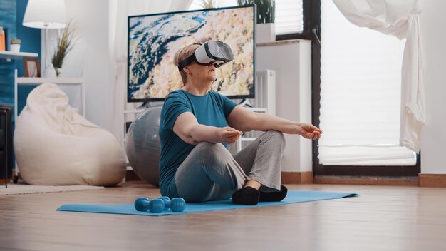 Mujer jubilada meditando en posición de loto con gafas vr en colchoneta de yoga. Pensionista usando auriculares de realidad virtual y haciendo meditación para relajarse. Persona mayor con gafas para meditar.