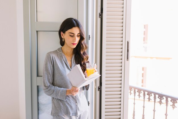 Mujer con jugo leyendo junto a la ventana