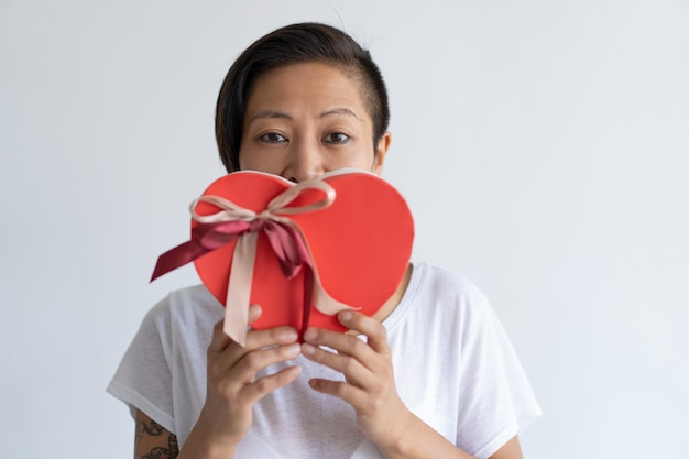 Mujer juguetona que sostiene la caja de regalo en forma de corazón delante de la boca