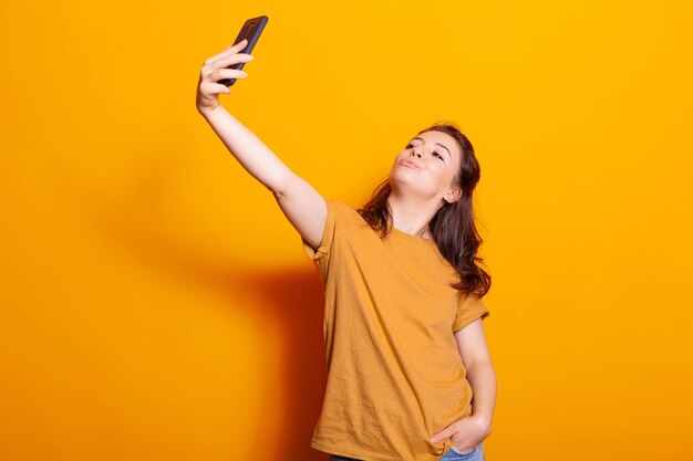 Mujer juguetona sosteniendo un teléfono móvil para tomar selfies en el estudio. Persona caucásica usando smartphone con tecnología, tomando fotos sobre fondo naranja. Adulto feliz con dispositivo moderno