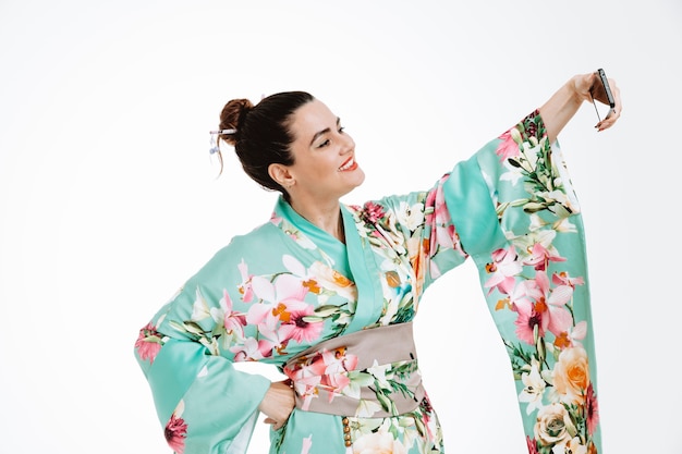 Mujer en kimono japonés tradicional feliz y positivo sonriendo alegremente haciendo selfie con smartphone en blanco