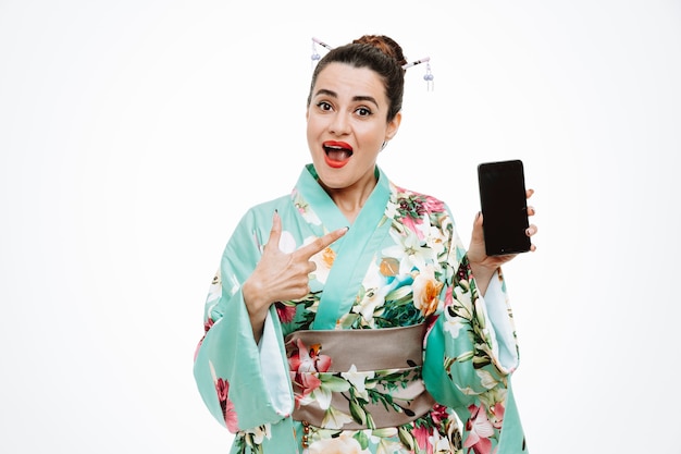 Mujer en kimono japonés tradicional sosteniendo smartphone apuntando con el dedo índice sonriendo confiado en blanco