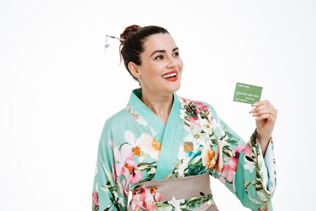 Mujer en kimono tradicional japonés con tarjeta de crédito mirando a un lado sonriendo confiado, feliz y positivo en blanco