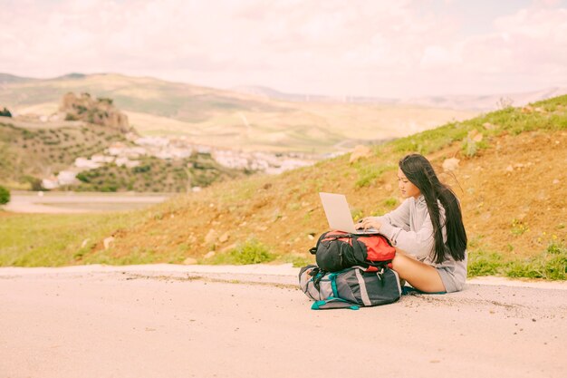 Mujer en el lado de la carretera y escribiendo en la computadora portátil en las mochilas