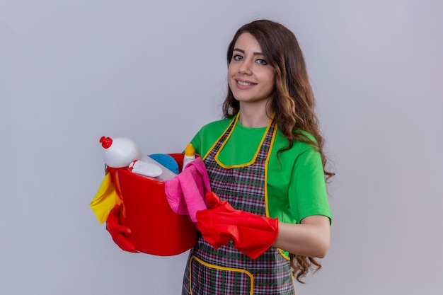 Mujer con largo cabello ondulado en delantal y guantes sosteniendo balde con herramientas de limpieza mostrando el pulgar hacia arriba sonriendo de pie amistoso