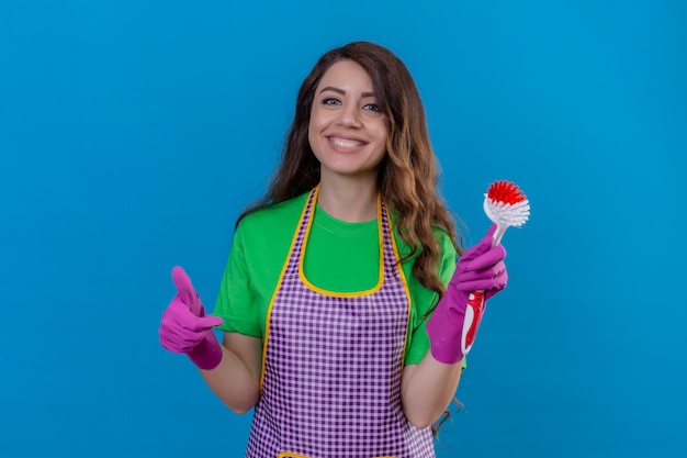 Foto gratuita mujer con largo cabello ondulado en delantal y guantes sosteniendo cepillo de fregar sonriendo alegremente mostrando el pulgar hacia arriba de pie en azul