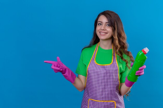 Mujer con largo cabello ondulado en delantal y guantes sosteniendo suministros de limpieza apuntando hacia el lado sonriendo alegremente de pie en azul