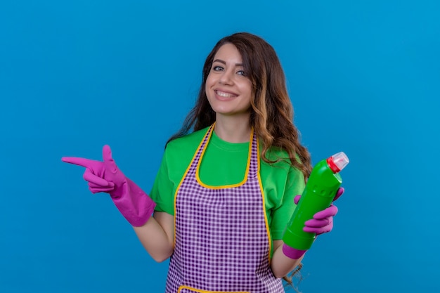 Mujer con largo cabello ondulado en delantal y guantes sosteniendo suministros de limpieza apuntando hacia el lado sonriendo alegremente de pie en azul