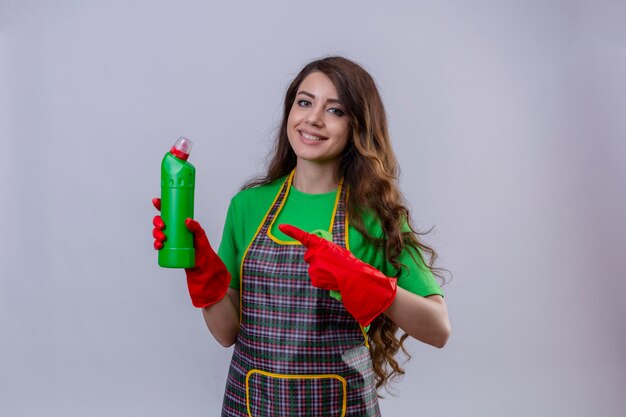 Mujer con largo cabello ondulado vistiendo delantal y guantes de goma apuntando con el dedo a una botella con productos de limpieza sonriendo de pie amable
