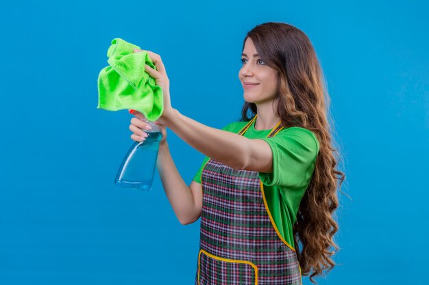 Mujer con largo cabello ondulado vistiendo delantal y guantes de goma sosteniendo una alfombra y spray de limpieza mirando confiada y positiva sonriendo lista para limpiar de pie en azul
