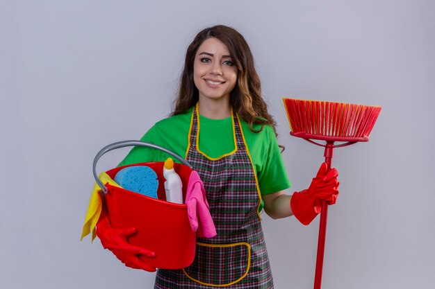 Mujer con largo cabello ondulado vistiendo delantal y guantes de goma sosteniendo un balde con herramientas de limpieza y un trapeador con cara feliz sonriendo alegre de pie