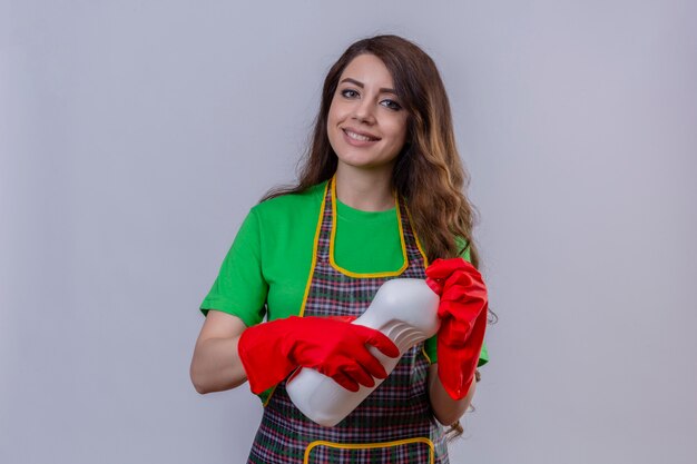 Mujer con largo cabello ondulado vistiendo delantal y guantes de goma sosteniendo una botella de productos de limpieza con aspecto positivo y feliz de pie y sonriendo