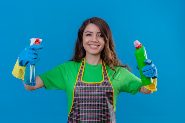 Mujer con largo cabello ondulado vistiendo delantal y guantes de goma sosteniendo botellas de productos de limpieza en manos levantadas sonriendo alegremente de pie sobre azul