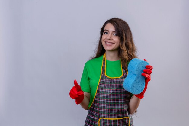 Mujer con largo cabello ondulado vistiendo delantal y guantes de goma sosteniendo una esponja mostrando los pulgares para arriba sonriendo amable de pie