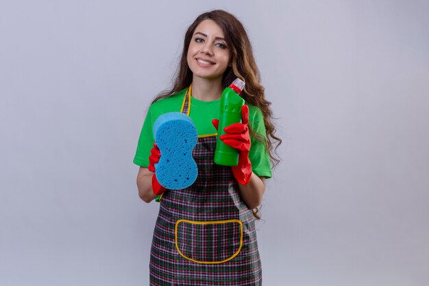 Mujer con largo cabello ondulado vistiendo delantal y guantes de goma sosteniendo una esponja y suministros de limpieza sonriendo amable de pie