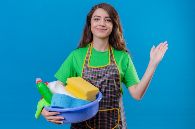 Mujer con largo cabello ondulado vistiendo delantal y guantes de goma sosteniendo lavabo con peaje de limpieza sonriendo saludando con la mano de pie en azul