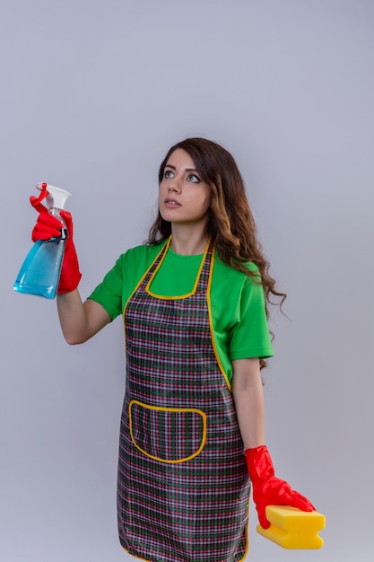 Foto gratuita mujer con largo cabello ondulado vistiendo delantal y guantes de goma sosteniendo spray de limpieza y una esponja mirando seguro listo para limpiar de pie
