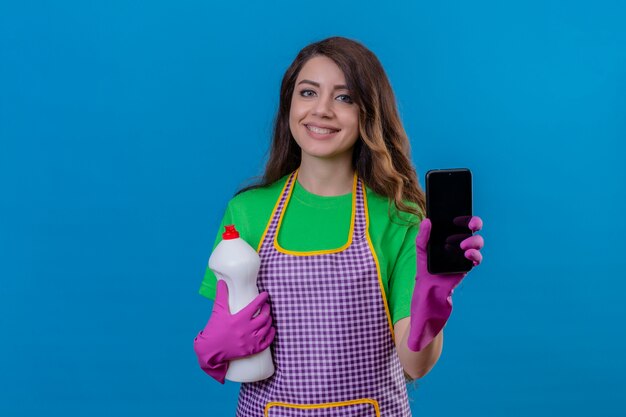 Mujer con largo cabello ondulado vistiendo delantal y guantes de goma sosteniendo suministros de limpieza mostrando teléfono móvil sonriendo amable de pie en azul