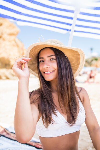 Mujer latina joven belleza en bikini y sombrero de paja sentado bajo la sombrilla en la playa cerca de la costa del mar.