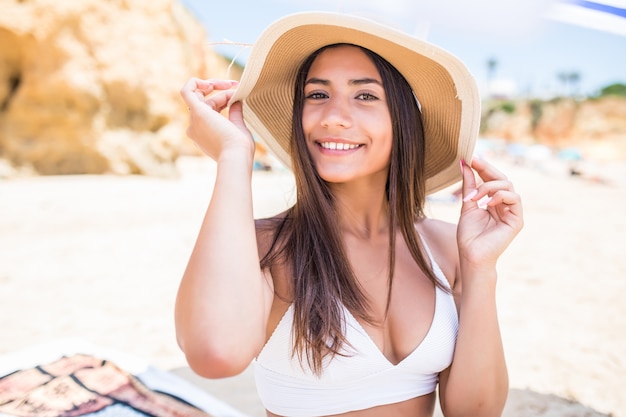 Mujer latina joven belleza en bikini y sombrero de paja sentado bajo la sombrilla en la playa cerca de la costa del mar.