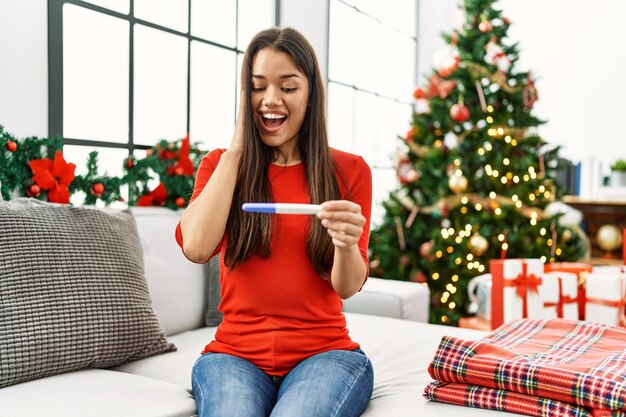 Mujer latina joven mirando prueba de embarazo sentada junto al árbol de navidad en casa