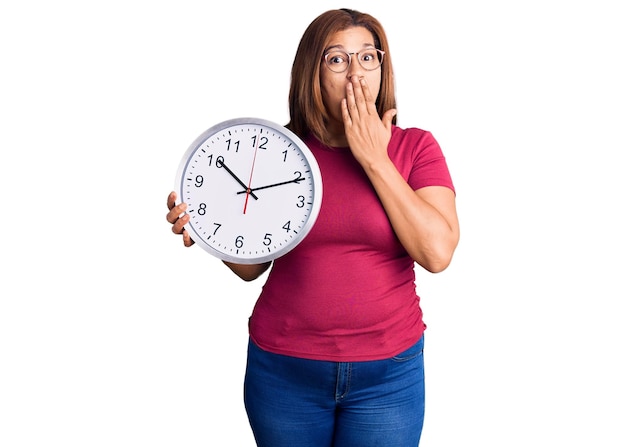 Foto gratuita mujer latina de mediana edad sosteniendo un gran reloj cubriendo la boca con la mano, sorprendida y temerosa de un error. expresión sorprendida