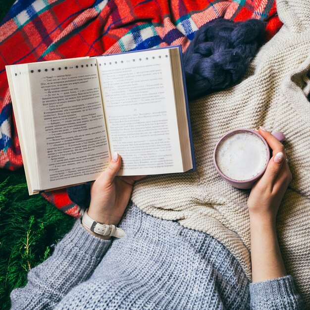 La mujer lee el libro que sostiene la taza de café y que miente bajo mantas coloridas