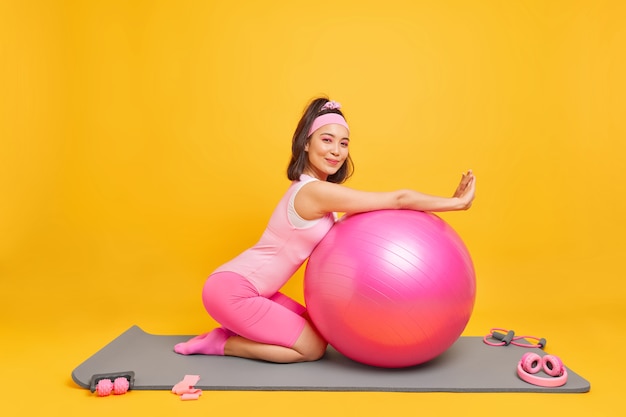 mujer lenas en fitness ball tiene expresión satisfecha vestida con ropa deportiva se toma un descanso después del entrenamiento en casa le gusta la gimnasia y el aeróbic posa en el tapete interior