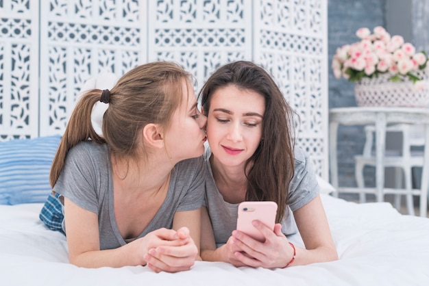 Mujer lesbiana joven que miente en cama que besa al polluelo de su novia usando el teléfono móvil