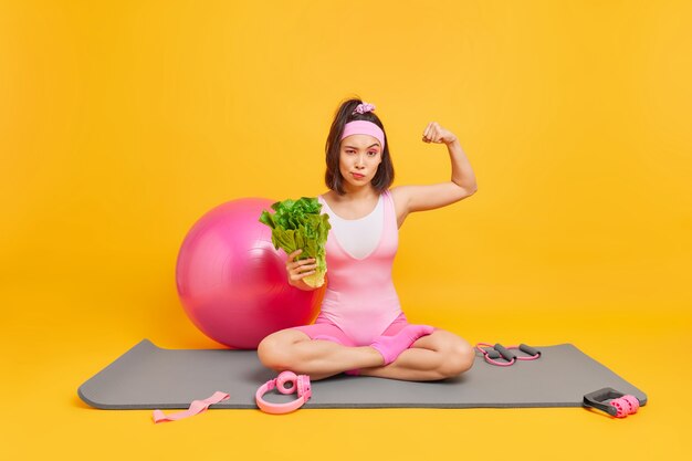 mujer levanta el brazo muestra los músculos después del entrenamiento mantiene una dieta saludable sostiene vegetales se sienta con las piernas cruzadas sobre la colchoneta de fitness con equipo deportivo alrededor. Estilo de vida saludable