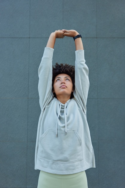 mujer levanta los brazos por encima de la cabeza se estira antes de que el entrenamiento se calienta hace ejercicios matutinos regulares plantea contra la pared gris mantiene un estilo de vida saludable