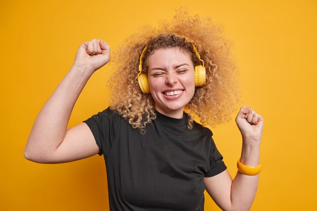 mujer levanta los brazos se siente alegre baila con el ritmo de la música escalofríos interiores usa auriculares estéreo camiseta negra casual