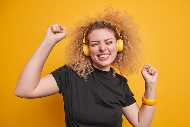 mujer levanta los brazos se siente alegre baila con el ritmo de la música escalofríos interiores usa auriculares estéreo camiseta negra casual