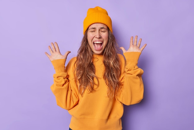 la mujer levanta las palmas exclama en voz alta mantiene la boca abierta viste un sombrero naranja y el saltador reacciona ante algo impresionante aislado en morado.