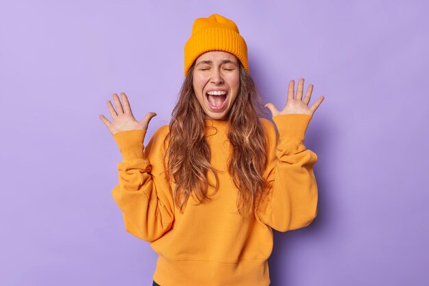 la mujer levanta las palmas exclama en voz alta mantiene la boca abierta viste un sombrero naranja y el saltador reacciona ante algo impresionante aislado en morado.