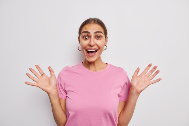 La mujer levanta las palmas se siente contenta sonríe alegremente reacciona ante noticias asombrosas vestida con una camiseta casual rosa sobre blanco