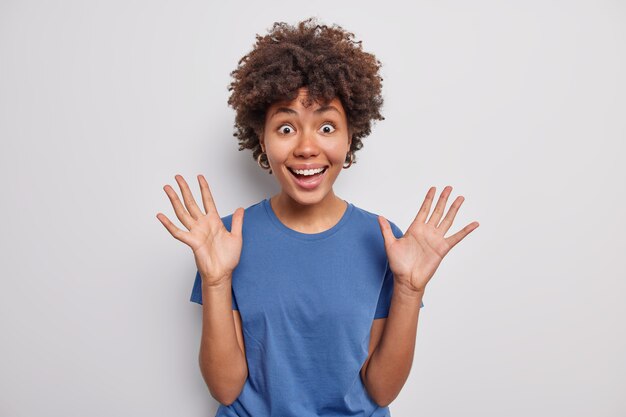 Mujer levanta palmas sonríe ampliamente reacciona con alegría ante algo gracioso expresa buenas emociones vestida casualmente plantea en blanco