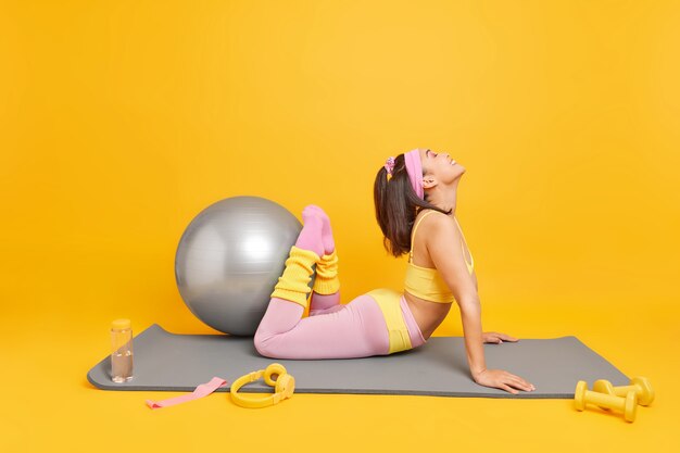 mujer levanta las piernas muestra su flexibilidad viste una camiseta recortada y leggings posa en una colchoneta de fitness hace ejercicios con pelota suiza se siente feliz aislado en amarillo