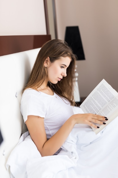 Mujer leyendo un libro y sonriendo mientras se acuesta en la cama matutina después de despertarse