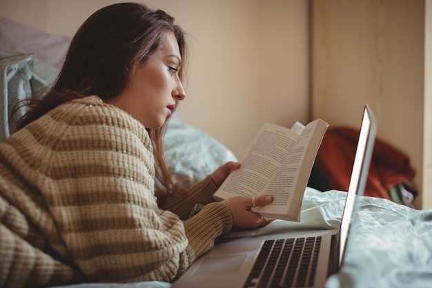 Mujer leyendo un libro y usando la computadora portátil en la cama