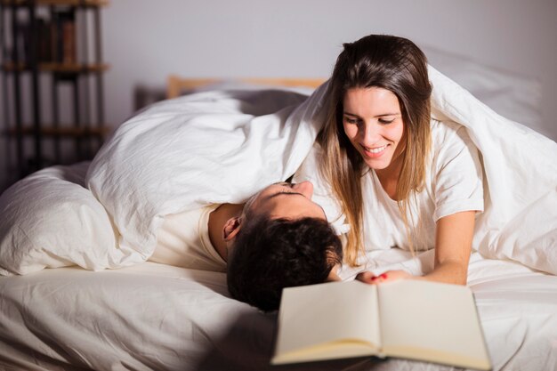 Mujer con libro cerca de hombre en edredón acostado en cama en la habitación