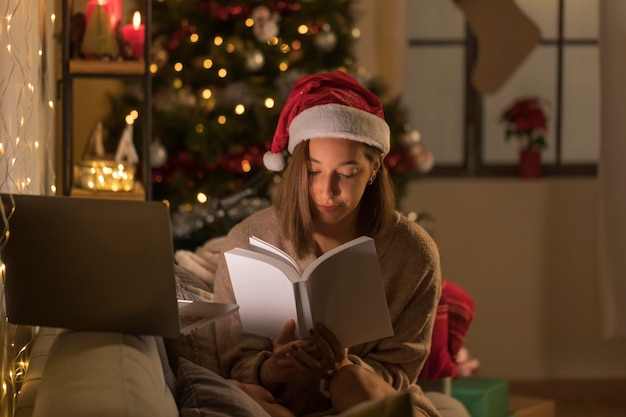 Mujer con libro de lectura de sombrero de santa delante de la computadora portátil